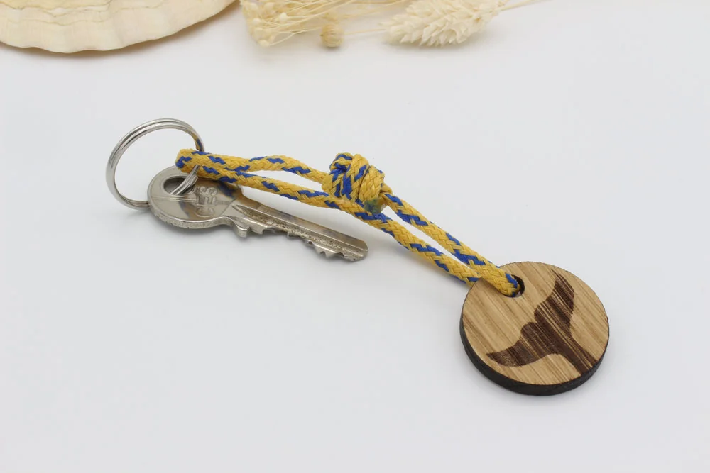 Gelb- blauer Schlüsselanhänger aus Geisternetz mit Holzscheibe und Walfluke. Am Edelstahl Schlüsselring befindet sich ein Schlüssel als Größenvergleich.